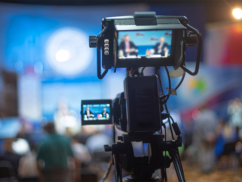 Video camera recording speakers at a live event.